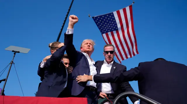 ۱۳/ ۷/ ۲۰۲۴، دونالد ترامپ، رئیس جمهور سابق، نامزد جمهوری خواه ریاست جمهوری، در حالی که توسط ماموران سرویس مخفی ایالات متحده محاصره شده است، هنگام خروج از صحنه در یک گردهمایی مبارزاتی، شنبه، ۱۳ ژوئیه ۲۰۲۴، در باتلر، ایالت پیا، اشاره می کند.