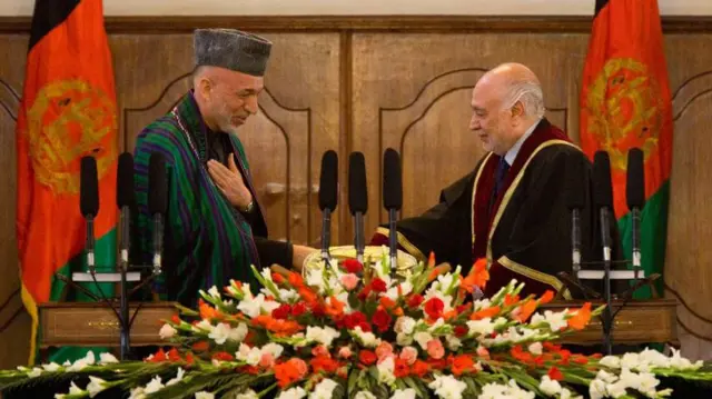 از راست به چب، عبدالسلام عظیمی و حامد کرزی رئیس جمهور پیشین افغانستان که در پشت هر دو پرچم دولت سابق افغانستان قرار دارد و در مقابل شان مایک و یک دسته بزرگ گل است.