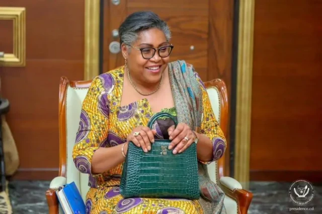 Judith Suminwa Tuluka porte un pagne à motif, elle a déposé sur ses deux genous un sac vert sur lequel elle a posé ses deux mains et sourit.