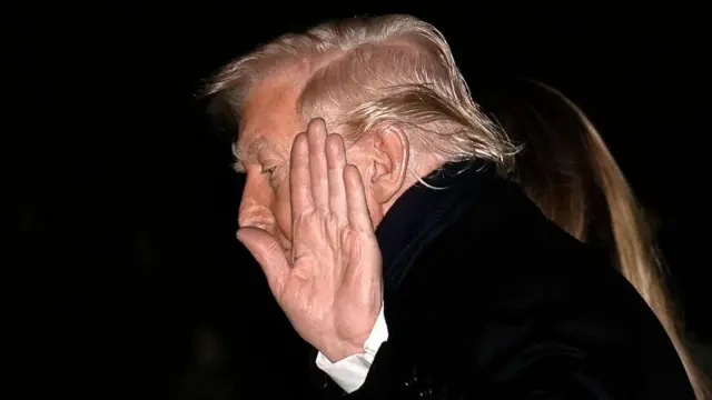 Дональд Трамп в профиль крупным планом, левая рука поднята в приветственном жесте, ладонь развернута к камере