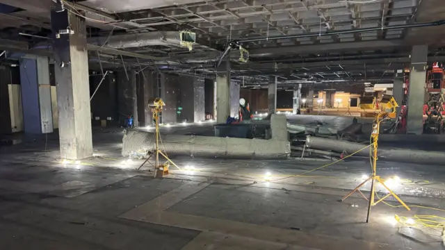 Un amplio espacio, similar a un almacén, está iluminado por pequeñas lámparas en el suelo. Niveles de burbuja y otros equipos de construcción se encuentran dispersos por el suelo de hormigón.