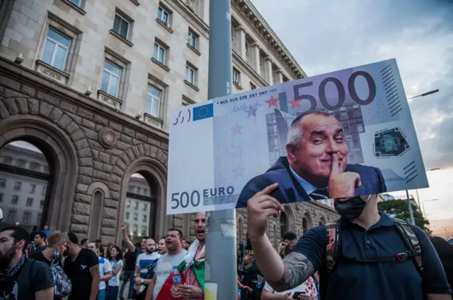Протестующий в Софии держит в руках плакат в виде купюры в 500 евро с улыбающимся премьером Бойко Борисовым. Снимок сделан 15 июля 2020 года.