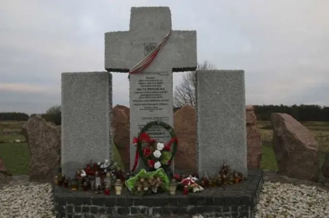 Пам’ятний меморіал у відкритому полі: у центрі кам’яний хрест із плитою з написом, перед ним вінки й букети квітів. Унизу розставлені лампадки. По боках — великі кам’яні брили; позаду видно поле й похмуре небо.