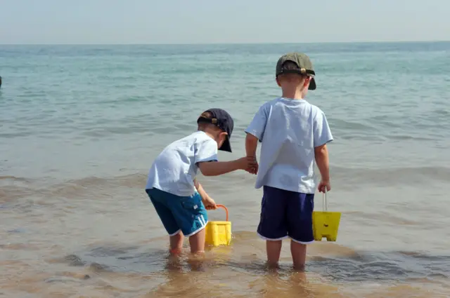 Dwoje dzieci ubranych w czapki, koszulki i spodenki stoi w płytkiej wodzie na plaży, każde trzymając żółte wiaderko