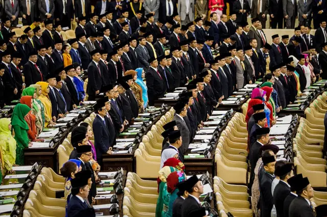 Pandangan dari jauh terlihat anggota DPR dengan pakaian formal berdiri dari tempat duduknya yang berbentuk melingkar.