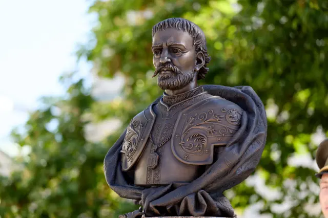 Бронзовий бюст Мазепи у латних обладунках на плечах, встановлений на відкритому повітрі на тлі зелених дерев.