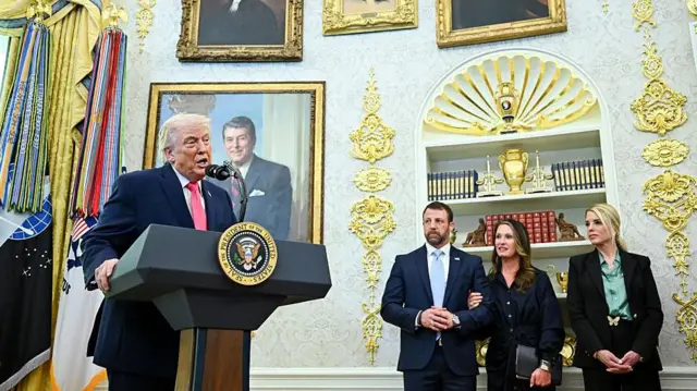 Trump kürsüde sağında bir erkek iki kadın ayakta bekliyor hepsi resmi giyimli. 