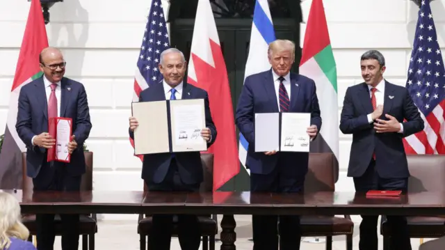 Ez-Zayani, Netanyahu, Trump ve Al Nahyan Beyaz Saray bahçesinde poz veriyor. Ellerinde İbrahim Anlaşmaları metni, arkalarında imza attıkları kahverengi masalar ve ülkelerinin bayrakları var.