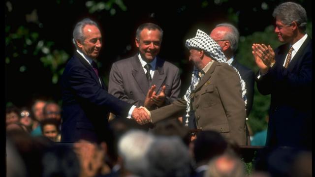 O líder da OLP, Yasser Arafat (à direita), apertando a mão do ministro das Relações Exteriores de Israel, Shimon Peres, na assinatura do Acordo de Oslo, com o presidente dos EUA, Bill Clinton (à direita), batendo palmas