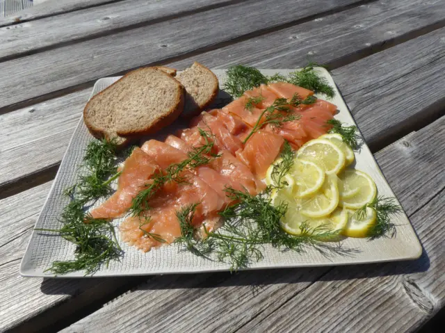 Un plato de pescado ahumado espolvoreado con eneldo con finas rodajas de limón y pan integral al lado.
