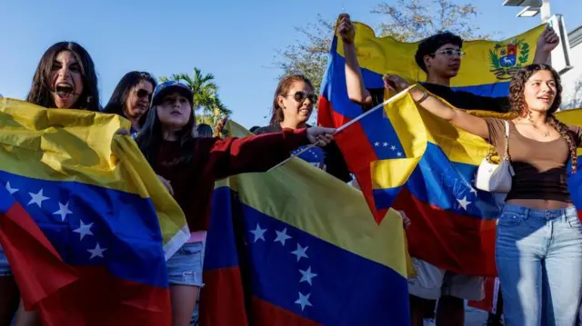 複数の若い男女が青空を背に、黄・青・赤のヴェネズエラ国旗を手に笑顔を浮かべたり叫んだりしている