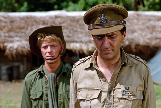 Двоє чоловіків у військовому одязі на тлі хатин із соломʼяними стріхами