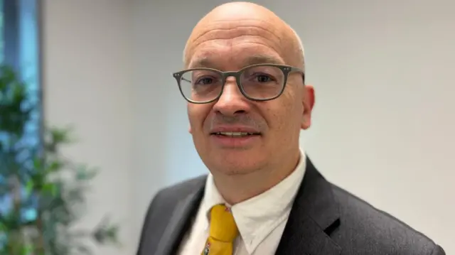 Ентоні одягнений у сірий костюм, білу сорочку та жовту краватку. Він носить окуляри в темній оправі та дивиться в камеру. Він стоїть в офісі з білим фоном та рослиною на задньому плані.