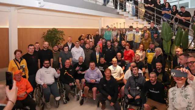 Групове фото з центру Superhumans: кількадесят ветеранів і працівників центру позують разом з принцом Гаррі. Принц, одягнений у куртку з назвою його Фонду "Ігри Нескорених" (Invictus Games Foundation), присів навпочіпки попереду