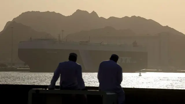 دو نفر در ساحل به یک کشتی در لنگرگاه نگاه می‌کنند