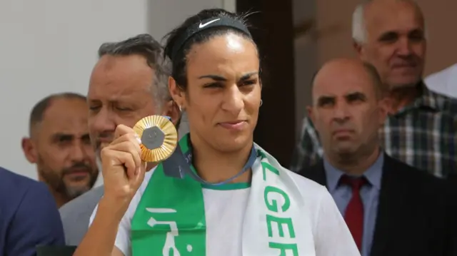 Imane Khelif sostiene una medalla. Hay varios hombres detrás de ella.