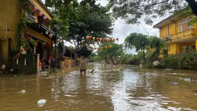 Foto of woman wey dey carry black bag for her head as she dey waka inside flood water.