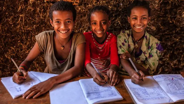 ፈገግ ያሉ ሦስት ታዳጊ ሴት ተማሪዎች ክፍል ውስጥ ደብተራቸው ተከፍቶ እስክሪብቶ ይዞ