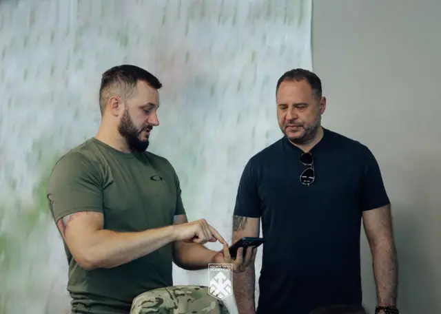 Двоє чоловіків стоять у приміщенні та дивляться на екран смартфона, який один із них тримає в руці. На передньому плані частково видно військове спорядження, а на зображенні є логотип підрозділу "Хартія".