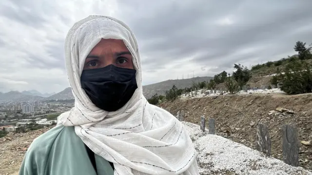 یوه ښځه چې په ماسک کې یې خوله پټه، سپين کیلې دارې ټېکری یې پر سر او پيکه زرغون رنګه کالي یې اغوستي دي، کامرې ته مخامخ ګوري. شا ته یې قبرونه او شناختې ښکاري او د کابل ښار ځينې ودانۍ هم برېښي.