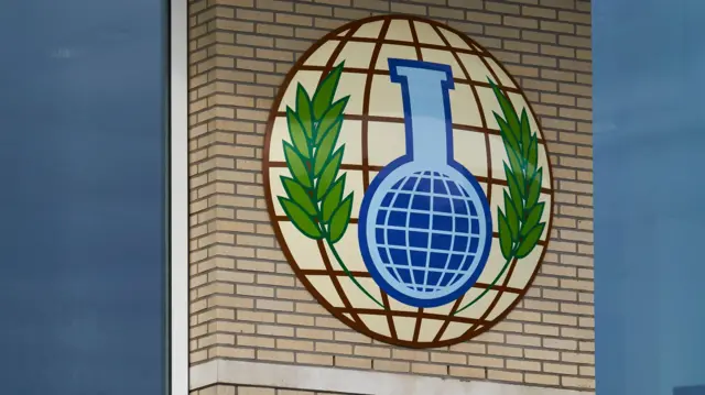 На кирпичной стене нарисован земной шар. На его фоне голубая колба в центре лаврового венка.