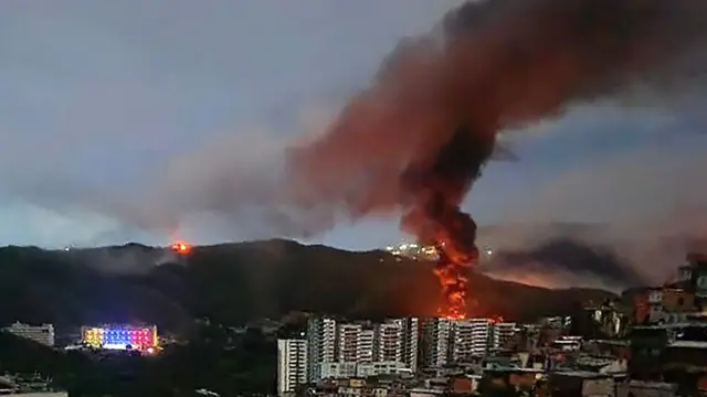 Un incendio en Fuerte Tiuna, el complejo militar más grande de Venezuela, se observa a la distancia tras una serie de explosiones en Caracas. Se ve una columna de humo elevándose hacia un cielo oscurecido, detrás de varios edificios altos y con una montaña al fondo.