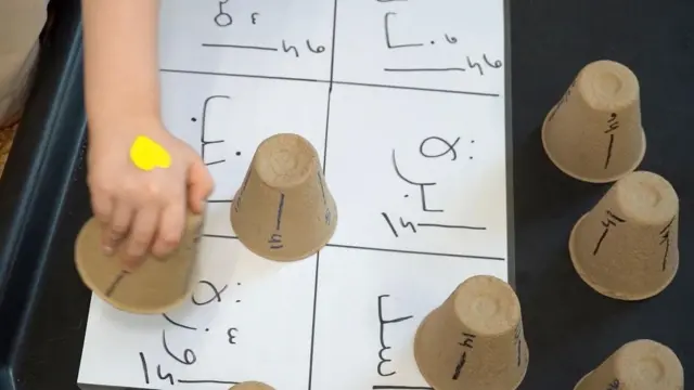 صورة تُظهر لوحة مكتوب عليها أحرفٌ باللغة العربية ويد طفل وهو يضع كوباً على كلمة إوزّة ضمن نشاطات حسّية لتعلّم اللغة العربية.