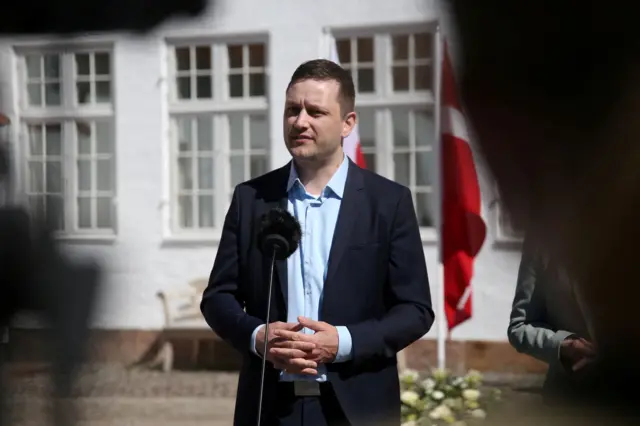 Jens-Frederik Nielsen, Greenland’s Prime Minister, wears a navy blue suit and a light blue shirt as he speaks to the press from behind a microphone. The Greenlandic and Danish flags are visible behind him.