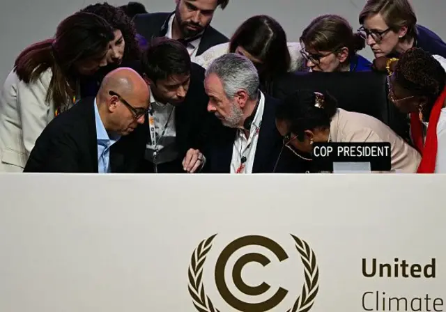 Dördü erkek beşi kadın bir grup resmi giyimli temsilci United Nations Climate yazdığı tahmin edilen bir kürsünün arkasında kafa kafaya müzakere ediyor.