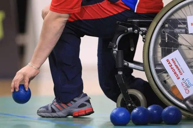 Jogadorbocha com bola na mão; no corte da imagem apenas as pernas, e o braço esquerdo do jogador são mostrados, alémbolas azul e parte da cadeirarodas.