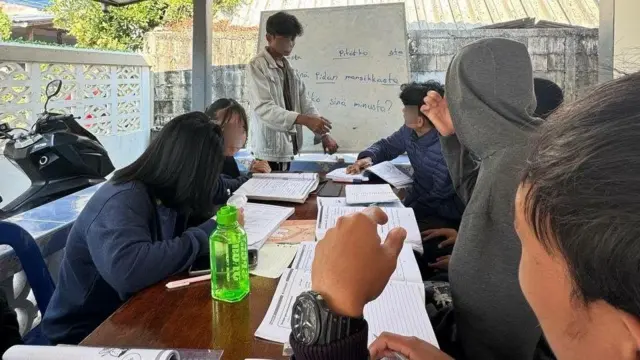 Một nhóm sinh viên đang ngồi quanh bàn với sách giáo khoa mở; trong khi một người đàn ông đang đứng trước bảng trắng trước nhóm