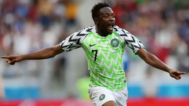 Ahmed Musa dans le maillot domicile vert fluo du Nigeria porté lors de la Coupe du monde 2018 en Russie. Le design comprend des chevrons blancs sur la poitrine verte et des chevrons noirs sur les manches blanches. Musa, visible des cuisses vers le haut, a les bras écartés et la bouche ouverte pour crier de fête alors qu'il court après avoir marqué un but. Une foule bondée est floue en arrière-plan