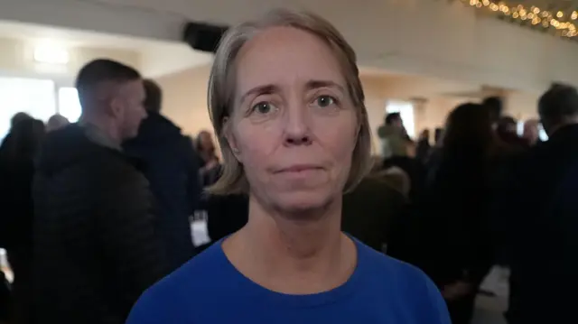 Sophie Marple, de pie en una sala durante una reunión. Mira directamente a la cámara y sonríe. Lleva una camiseta azul.