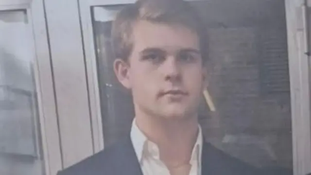 Achille Osvaldo Giovanni Barosi ma na sobie koszulę z rozpiętym kołnierzykiem i marynarkę.