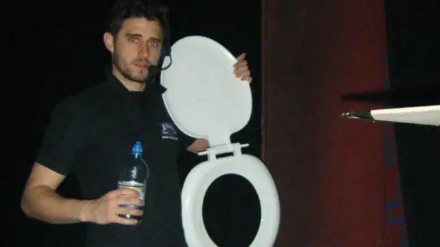 Ben Parsley, sosteniendo una botella de agua, vestido completamente de negro, con auriculares y sosteniendo un asiento de inodoro blanco, está detrás del escenario en un teatro.