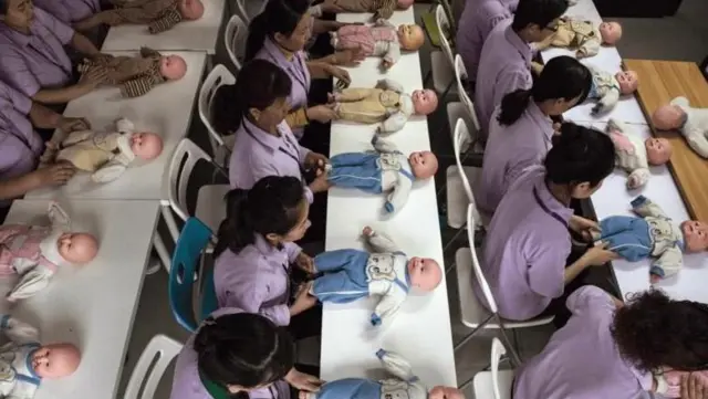 نمایی از بالا، افراد پشت میزها به ردیف کنار هم نشسته‌اند و جلوی هر یک از آنها روی میز، یک عروسک یک شکل قرار گرفته است