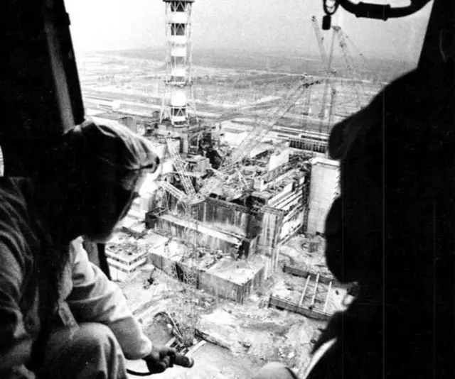 Чорно-біле фото, зняте з повітря: двоє людей у захисному одязі сидять у відкритих дверях вертольота й дивляться вниз на зруйнований промисловий комплекс з високою трубою, кранами та пошкодженими будівлями. Унизу — масштабні руйнування й техногенний ландшафт.
