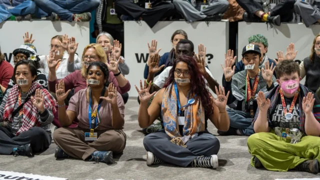 Des personnes de différentes ethnies assises avec du ruban adhésif sur la bouche portant le message « payez ».
