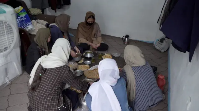 सिर पर स्कार्फ बांधे छह महिलाएं खाने की मेज के चारों ओर फर्श पर पैर मोड़कर बैठी हैं.