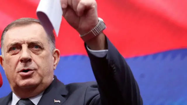 Милорад Додик у оделу и подигнутом руком у којој држи беле папире