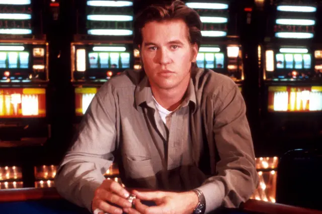 Val Kilmer is seated at a casino table with hands resting on the edge, holding a small object. He is wearing a light-coloured button-up shirt over a white undershirt and a wristwatch. In the background, there are several brightly lit slot machines arranged in rows, creating a colourful and vibrant setting typical of a casino environment. The photo is a still from his 1989 film, Kill Me Again.