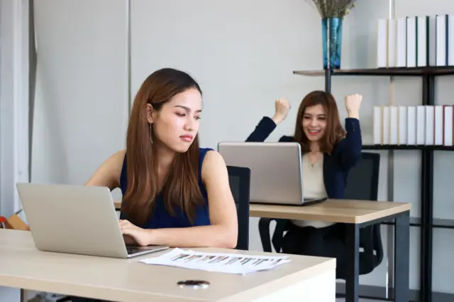 Em escritório, mulher demonstra olhar invejoso enquanto amiga, na mesa atrás, comemora 