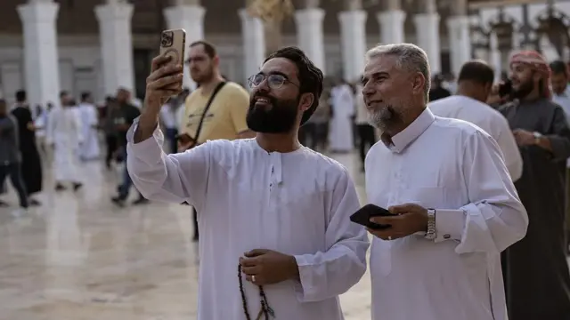 Two men wearing white jalamia holding phones, one dey use im phone take selfie as dem dey both smile.