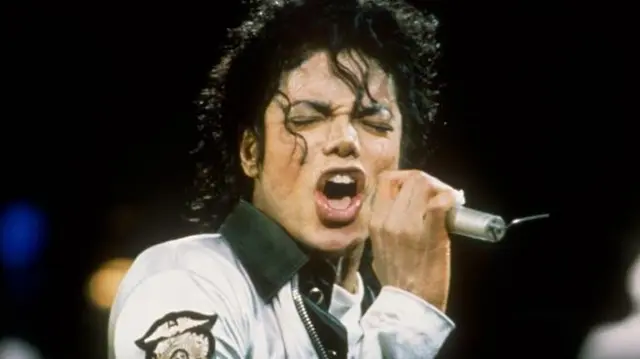 Amerykański piosenkarz Michael Jackson podczas koncertu w Londynie w 1998 roku. Ma na sobie biało-czarną kurtkę i śpiewa do mikrofonu z zamkniętymi oczami.