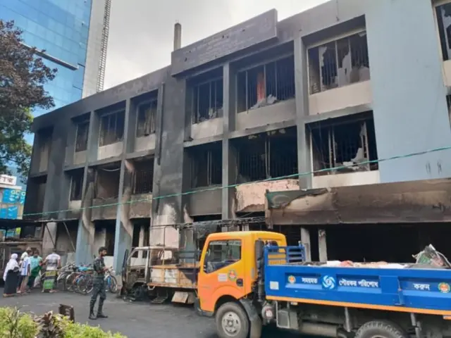আগুনে ভস্মীভূত মিরপুর থানা। সামনে দুটি পিকআপ ট্রাক ও বাইসাইকেল। কয়েকজন মানুষ দাঁড়িয়ে আছেন