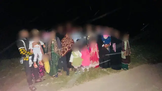 রাতের অন্ধকারে কয়েকজন নারী-পুরুষ ও শিশু দাঁড়িয়ে আছে যাদের মুখ ঝাপসা করে দেওয়া আছে