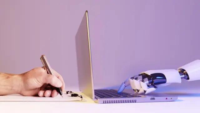 صورة يد تحمل قلماً وتكتب على الورق مقابل يد آلية تكتب على جهاز كمبيوتر.