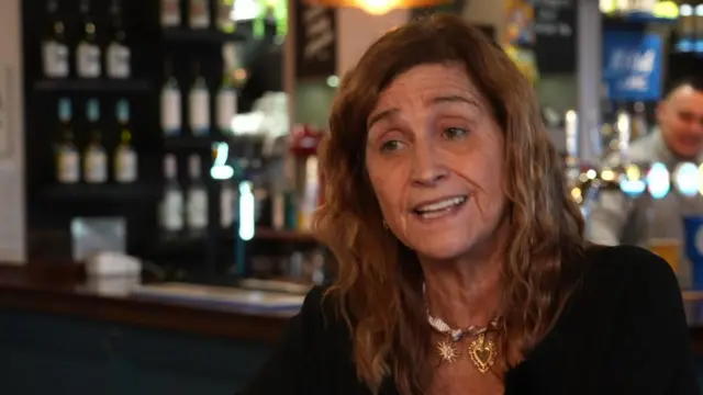 Ellie Pitt, con el pelo hasta los hombros y una blusa negra y un collar. Al fondo, el interior de un pub.