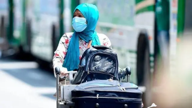 विमानस्थलमा ट्रलीमा झोला र सुट्केश राखेर हिँडिरहेकी एक महिला। उनले मास्क लगाएकी छन् र निलो सलले टाउको ढाकेकी छन्। पृष्ठभूमीमा हरियो रङ्गको बस देख्न सकिन्छ