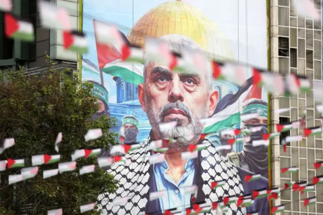 ما نراه صورة ضخمة ليحيى السنوار بنظرة ثاقبة وقد رفعت في أحد الشوارع المزينة باعلام فلسطينية فوق المارة وفي خلفية الصورة مسجد قبة الصخرة ومقاتلو كتائب القسام